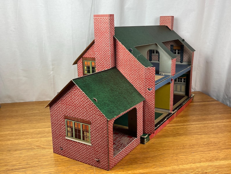 May include: A vintage-style dollhouse featuring a red brick exterior pattern, a dark green gabled roof, and two prominent chimneys. The structure is open on one side, revealing multiple interior rooms with colored walls and printed window details.