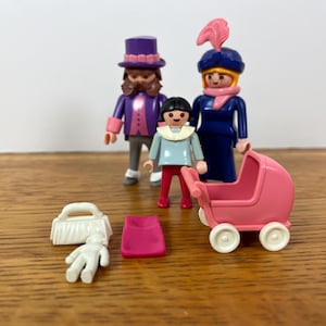 Könnte beinhalten: Eine Sammlung von Playmobil-Figuren und Zubehör. Das Set enthält eine dreiköpfige Familie, einen rosa Kinderwagen, eine weiße Handtasche, eine rosa Decke und eine kleine weiße Babyfigur. Die Figuren tragen verschiedene Outfits, darunter einen lila Zylinder und ein blaues Kleid.