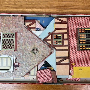 May include: A cardboard model house kit in a box, featuring flat pieces with printed stone, red brick, and timber-frame patterns. The set includes various wall sections with illustrated windows, a door, and a circular architectural detail.