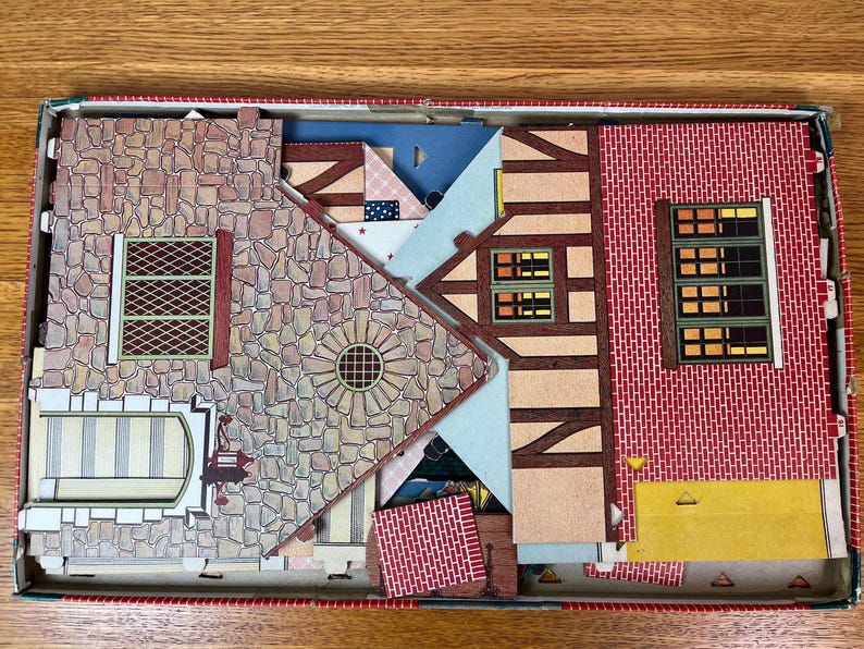 May include: A cardboard model house kit in a box, featuring flat pieces with printed stone, red brick, and timber-frame patterns. The set includes various wall sections with illustrated windows, a door, and a circular architectural detail.
