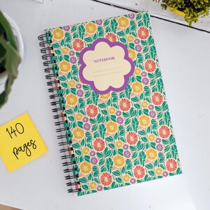 Könnte beinhalten: Ein spiralgebundenes Notizbuch mit einem floralen Muster auf dem Einband. Der Einband zeigt eine violette Blütenform mit dem Wort "NOTEBOOK" darin. Das Notizbuch hat 140 Seiten.
