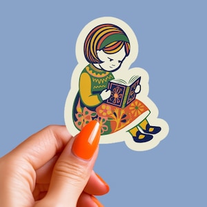 May include: A sticker featuring a seated figure reading a book. The figure is wearing a green and yellow sweater, a floral skirt, and black shoes. The book has a decorative cover. The sticker has a light blue background.