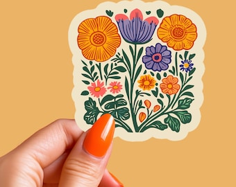 May include: A floral sticker featuring a variety of colorful flowers in shades of orange, purple, pink, and yellow. The sticker has a cream-colored background and is held by a hand with orange nail polish.