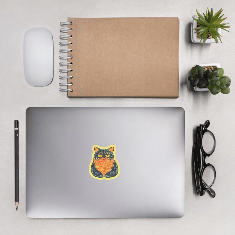 May include: A sticker featuring a cartoon cat with orange fur and black eyes. The cat is sitting with its paws tucked under its body. The sticker is on a light gray laptop.