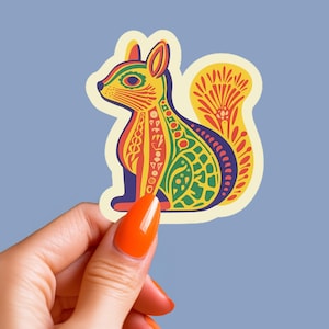 May include: A colorful sticker featuring a stylized squirrel design. The squirrel is depicted in shades of yellow, red, green, and purple, with intricate patterns. The sticker is held against a light blue background.