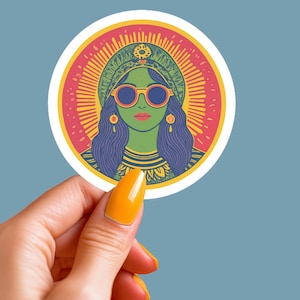Peut inclure: Un autocollant rond avec une illustration colorée d'une femme portant des lunettes de soleil et une couronne. L'arrière-plan est un soleil éclatant rouge et jaune.