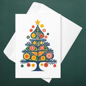 May include: A white greeting card with a stylized Christmas tree design. The tree is navy blue with yellow, orange, and red ornaments and a yellow star on top. The card is inside a white envelope, set against a dark green background.