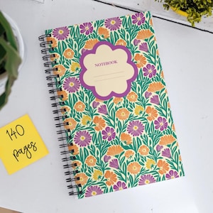 Könnte beinhalten: Ein spiralgebundenes Notizbuch mit einem Blumendesign mit lila, orangefarbenen und grünen Blumen auf cremefarbenem Hintergrund. Der Einband hat ein lilafarbenes Etikett mit der Aufschrift "NOTEBOOK".