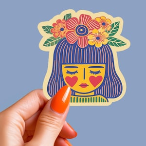 Pode incluir: Um autocolante com uma ilustração estilizada do rosto de uma mulher com cabelo azul e uma coroa de flores. A mulher tem bochechas em forma de coração e olhos fechados. O autocolante é segurado por uma mão com verniz de unhas laranja sobre um fundo azul claro.