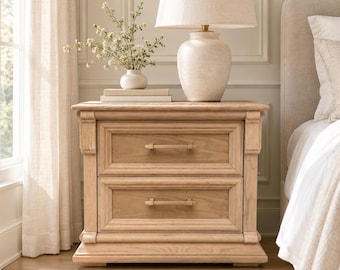 May include: A light oak wood bedside table featuring two drawers with horizontal metal handles. The bedside table is topped with a ceramic table lamp with a beige fabric shade and a small white vase containing green sprigs.