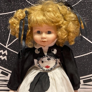 May include: A porcelain doll with curly blonde hair tied with black ribbons, blue eyes, and a pale complexion. The doll wears a black and white dress featuring a circular patch on the chest with a stitched face design and a red diamond shape.