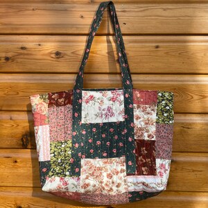 May include: A quilted tote bag featuring a patchwork design of various floral patterns in shades of pink, burgundy, green, and cream. The bag includes two long shoulder straps made from a dark green fabric with a small floral print pattern.
