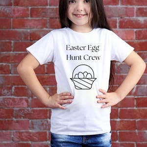 Peut inclure: Un t-shirt blanc avec le texte "Easter Egg Hunt Crew" et une illustration en noir et blanc d'un panier avec des œufs.