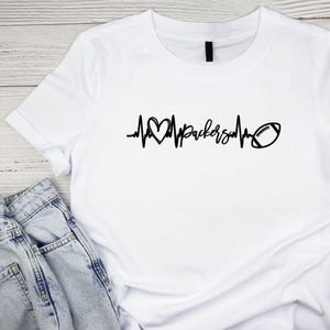 May include: White t-shirt with a black graphic design. The design features a heartbeat line with a heart, the word "Packers", and a football. The shirt is paired with a pair of blue jeans.