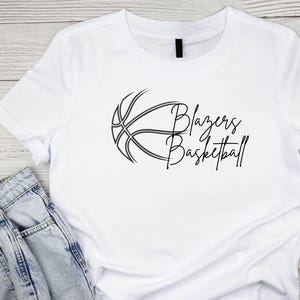 May include: White t-shirt with a black basketball graphic and the text "Blazers Basketball" in a cursive font.