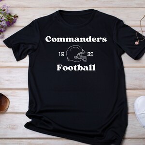 May include: Black t-shirt with white text that reads "Commanders Football" with a football helmet graphic and the numbers 19 and 32.