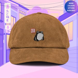 Op de afbeelding: Een bruine corduroy baseballcap met een geborduurde kikker en Japanse karakters. De kikker is zwart met een witte buik en zit op een rood vierkant met witte karakters.