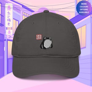 Könnte beinhalten: Eine dunkelgraue Baseballkappe mit einer gestickten schwarzen Frosch und einem roten Kanji-Zeichen. Das Kanji-Zeichen ist "蛙", was im Japanischen "Frosch" bedeutet.