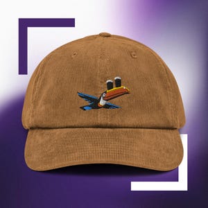 以下が含まれることがあります： 2つのビールグラスを持った刺繍のオニオオハシが描かれた、茶色のコーデュロイ製のベースボールキャップです。