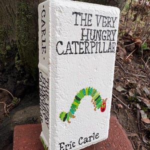 May include: A white, rectangular decorative piece resembling a book, featuring the title "THE VERY HUNGRY CATERPILLAR" in black lettering. A colorful caterpillar illustration is on the front, with the author's name, Eric Carle, below. The book is placed on a red brick.