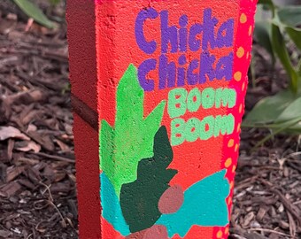 May include: A red brick painted with green, brown, and blue accents. The brick has the words "Chick Chick Boom Boom" written in purple letters.