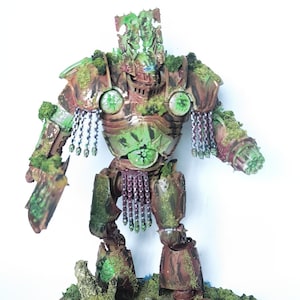 May include: A detailed miniature robot figure, primarily brown and green, with intricate armor and detailing. The figure is adorned with moss-like textures and hanging chains, standing on a base with miniature foliage and rocks. The robot has a menacing appearance.