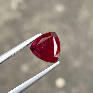 Puede incluir: Una gema de rubí facetada en forma triangular. El rubí es de un color rojo intenso y profundo, sostenido por unas pinzas plateadas. La gema tiene una superficie pulida y bordes afilados, reflejando la luz.