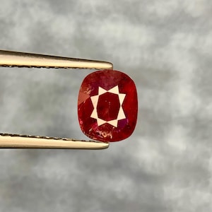 May include: A cushion-cut red gemstone held by metal tweezers. The stone features a distinct, bright white star-shaped reflection in its center, contrasting with the deep, translucent red body of the faceted gem, showcasing internal clarity and color.