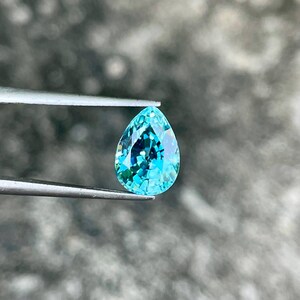 May include: A teardrop-shaped blue gemstone with a pearlescent finish. The gemstone is held by tweezers against a blurred background.