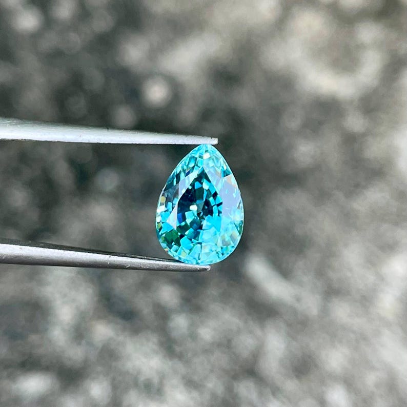 May include: A teardrop-shaped blue gemstone with a pearlescent finish. The gemstone is held by tweezers against a blurred background.