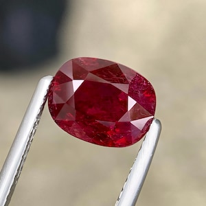 Könnte beinhalten: Ein tiefroter Edelstein im Kissenschliff mit facettierter Oberfläche, gehalten von einer silberfarbenen Metallpinzette. Der Stein zeigt einen satten, kräftigen Karmesinton mit innerer Klarheit und lichtreflektierenden Facetten.