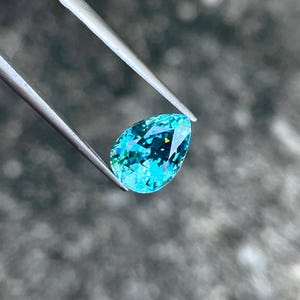 May include: A pear-shaped teal gemstone with a light blue hue and a hint of green. The gemstone is held by tweezers against a blurred background.