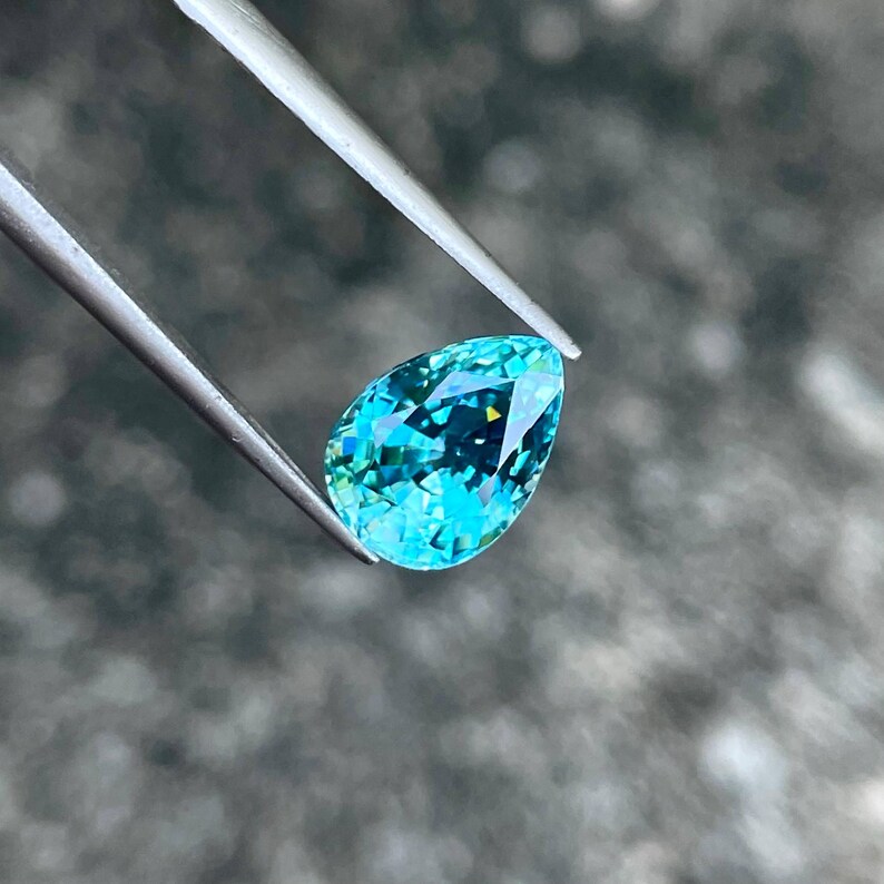 May include: A pear-shaped teal gemstone with a light blue hue and a hint of green. The gemstone is held by tweezers against a blurred background.