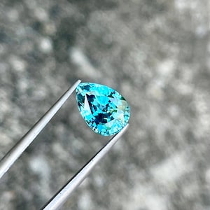 May include: A pear-shaped teal gemstone with a light blue hue, held in tweezers against a gray background.