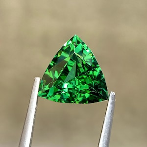 Könnte beinhalten: Ein leuchtend grüner Edelstein im Dreiecksschliff mit zahlreichen Facetten, die das Licht reflektieren. Der Edelstein wird von einer silberfarbenen Metallpinzette gehalten, was seine Transparenz und die tiefe, gesättigte grüne Farbe hervorhebt.