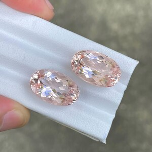 May include: Two oval-shaped, light pink gemstones with a faceted cut. The gemstones are displayed on a white surface.