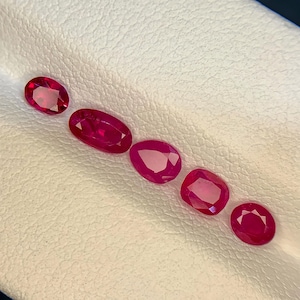 May include: A collection of six oval and round-cut rubies. The gemstones are a vibrant red color and are arranged in a diagonal line on a white surface. The rubies vary slightly in size and shape.
