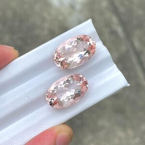 May include: Two oval-shaped, pale pink gemstones with a faceted cut. The gemstones are displayed on a white surface.