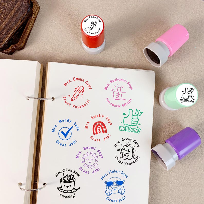 K&ouml;nnte beinhalten: Eine Sammlung farbenfroher Stempel mit verschiedenen Designs und Phrasen wie "Vertraue dir selbst!", "Tolle Arbeit!" und "Gut gemacht!". Die Stempel sind in Rot, Pink, Gr&uuml;n und Lila gehalten und hinterlassen wei&szlig;e Abdr&uuml;cke auf wei&szlig;em Papier.