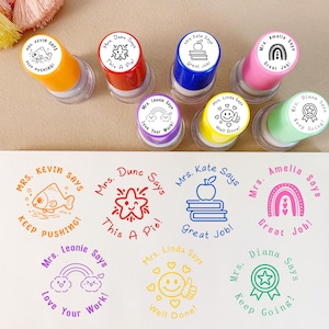 K&ouml;nnte beinhalten: Eine Sammlung farbenfroher Selbstf&auml;rbestempel mit verschiedenen Designs und Phrasen. Die Stempel zeigen Bilder von Fisch, Stern, Apfel, Regenbogen und Smiley, mit S&auml;tzen wie "Keep Pushing!" und "Great Job!"