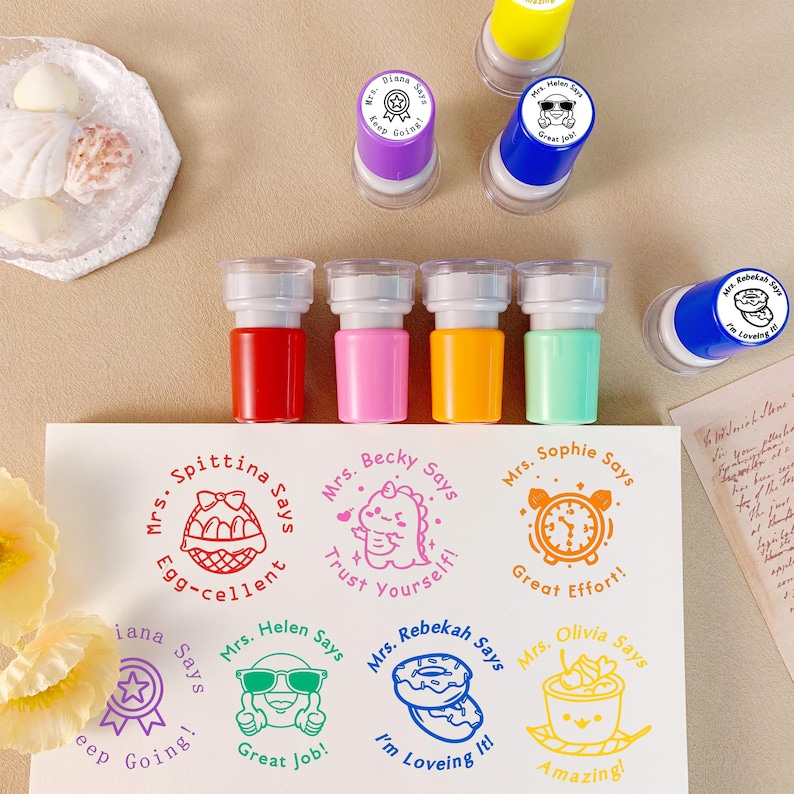 K&ouml;nnte beinhalten: Eine Sammlung farbenfroher Selbstf&auml;rbestempel mit verschiedenen Designs und Phrasen. Die Stempel enthalten S&auml;tze wie &bdquo;Egg-cellent&ldquo;, &bdquo;Great Job!&ldquo; und &bdquo;Amazing!&ldquo;. Die Stempel sind in Rot, Pink, Orange, Gr&uuml;n, Gelb und Blau.