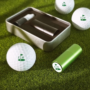 May include: A collection of golf balls and a green golf accessory are displayed on artificial grass. The golf balls feature a green flag logo with the letters "EM". A silver tin holds two cylindrical items. The green accessory also has the "EM" logo.