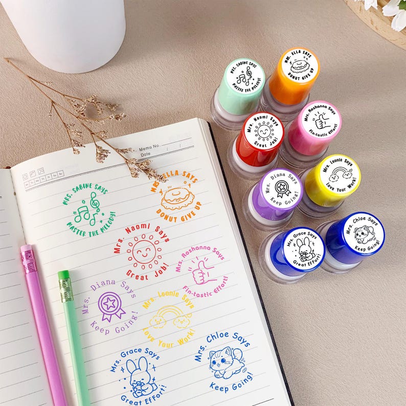 K&ouml;nnte beinhalten: Eine Sammlung bunter Lehrerstempel mit verschiedenen Designs und Phrasen, darunter eine Musiknote, ein Donut, eine Sonne, ein Daumen hoch, ein Regenbogen, ein Stern, ein Hase und eine Katze. Die Stempel befinden sich auf einem Notizbuch mit passenden gestempelten Bildern.