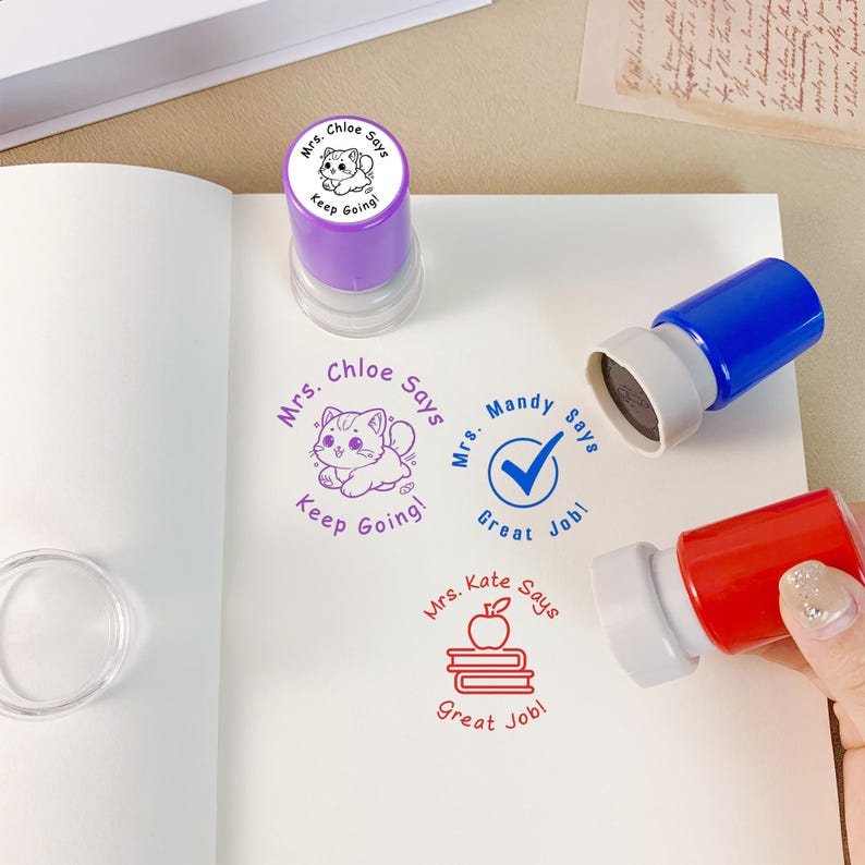 K&ouml;nnte beinhalten: Eine Sammlung farbenfroher Lehrerstempel mit verschiedenen Designs und Phrasen. Die Stempel umfassen einen lila Stempel mit einer Katzenzeichnung und den Worten "Mrs. Chloe Says Keep Going!" und einen blauen Stempel mit einem H&auml;kchen.