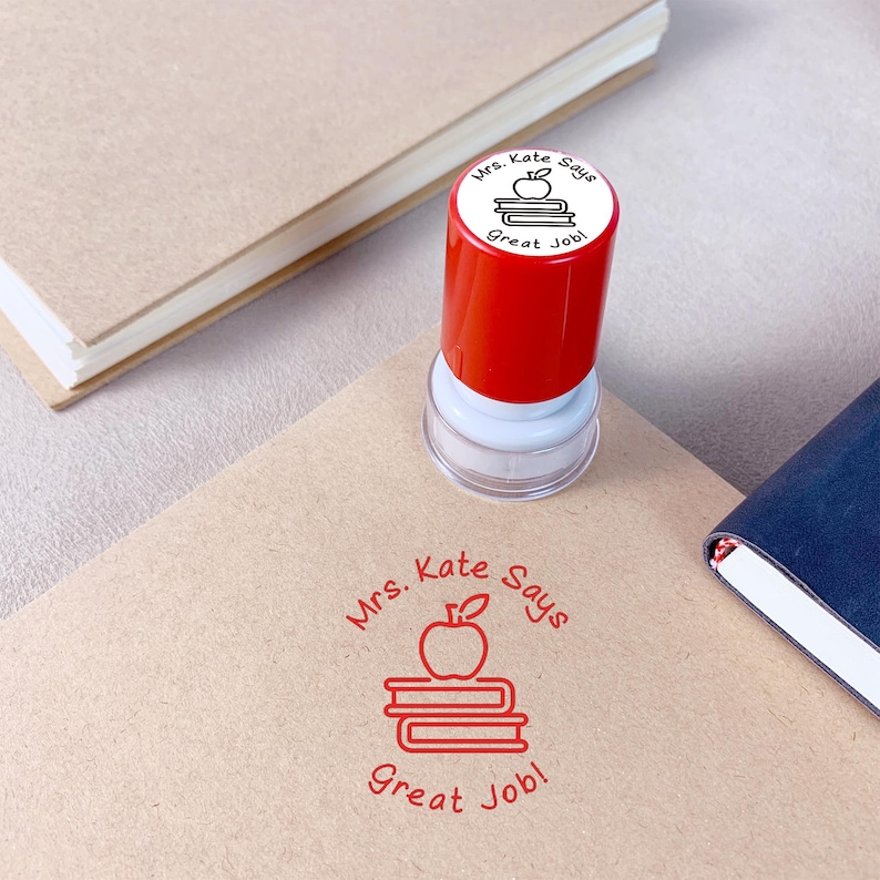 K&ouml;nnte beinhalten: Ein rot-wei&szlig;er Stempel mit dem Text "Mrs. Kate Says Great Job!" und einem Apfel auf B&uuml;chern. Der Stempel befindet sich auf einer beigefarbenen Oberfl&auml;che mit einem passenden Stempelbild.