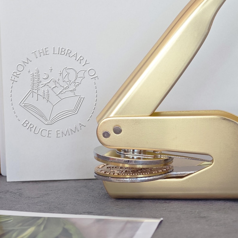May include: A gold-colored self-inking stamp with a round design featuring a book, trees, a crescent moon, and a bat. The text "From the Library of Bruce Emma" is inscribed around the design.