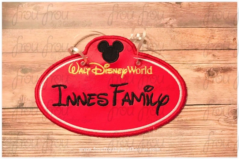 May include: Red oval-shaped name tag with white trim, featuring a black Mickey Mouse head at the top, the text "Walt Disney World" in yellow, and the text "Innes Family" in black. The name tag is on a wood background.