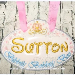 Op de afbeelding: Een witte ovale deurhanger met een roze lint, een ontwerp van een Assepoesterkoets en de tekst "Sutton Bibbidi Bobbidi Boo" in gouden en blauwe letters. De tekst "www.froufroubyheathersue.com" staat onderaan gedrukt.