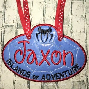 Op de afbeelding: Blauw en rood geborduurd naamschild met een spinnenwebontwerp en de naam "Jaxon Islands of Adventure" in rode letters.