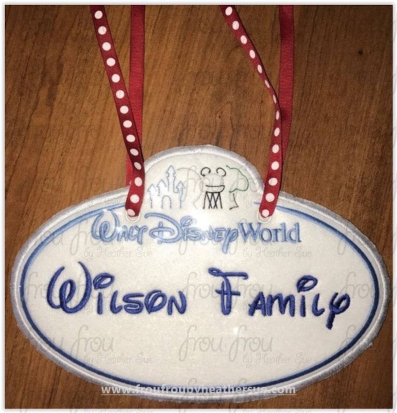 May include: White oval-shaped name tag with blue embroidered text that reads "Walt Disney World" and "Wilson Family". The tag has a red ribbon with white polka dots attached to the top.
